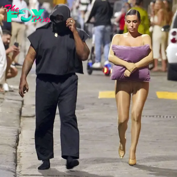 Bianca Censori, holding a pillow to her chest, walking with Kanye West on a street