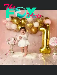 This may contain: a baby girl standing in front of a number one balloon with flowers and balloons around her