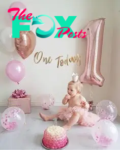 This may contain: a baby girl sitting on the floor in front of a pink cake with one year balloons