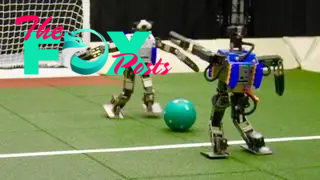 Humanoid robots stand either side of a football in front of a goal post.