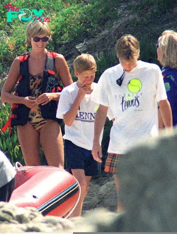 Princess Diana in a swimsuit, apparently tiger print, wearing sunglasses and a life vest. Her sons are in front of her, Prince Harry in a white t-shirt and blue shorts and Prince William looking down in a white t-shirt marked "Totally tennis" with a large tennis ball logo. They are on rocks and in front of them appears to be an inflatable boat.