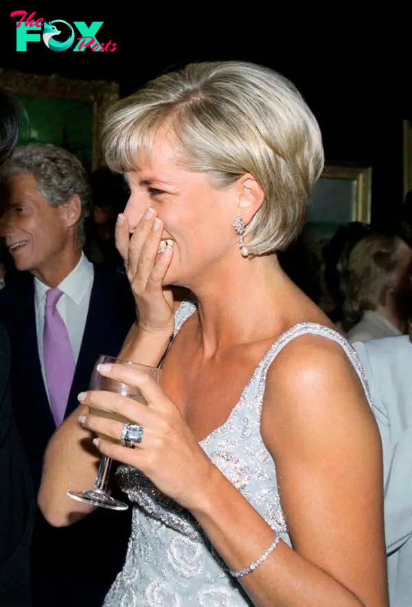 Princess Diana covers her mouth with her hand laughing while wearing a party dress and holding a glass in her left hand, making an aquamarine ring visible.