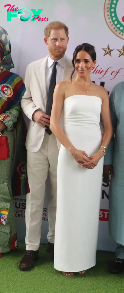 Meghan Markle in a long strapless white dress wearing a cross necklace, with Prince harry in a light tan suit standing behind her