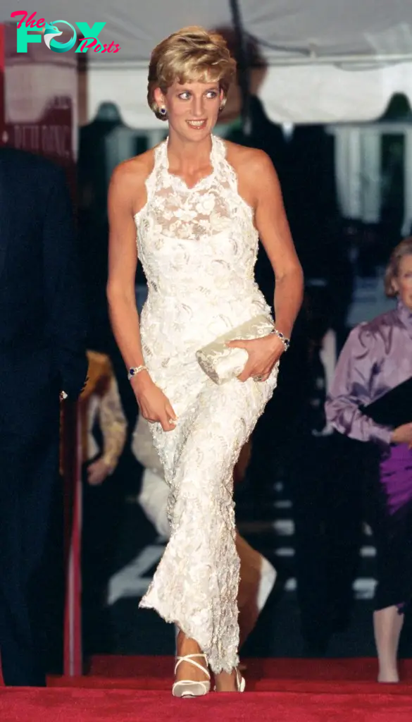 Diana in a long white dress with bare shoulders and apparent halter top, clutching a white purse. She is wearing two emerald bracelets.