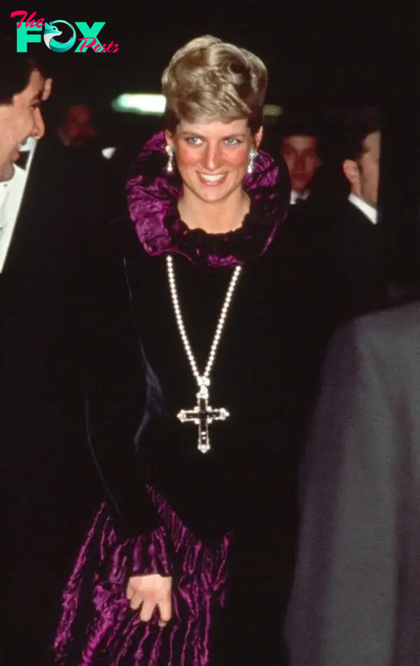 Princess Diana smiling and wearing a long black dress with ruffled purple collar and ruffled collars and lower half. She is wearing a large jeweled pectoral cross on a rope of pearls.