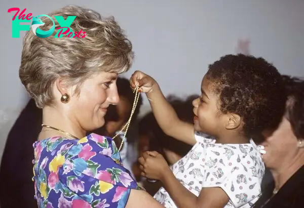 Princess Diana holds a toddler in her arms who lifts a necklace into her hands.