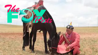 A man in a pink robe and black hat sits on the ground next to a saddled horse on a grassy plain.