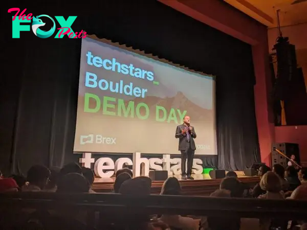 Gov. Jared Polis talks to a crowd while standing on a stage. Behind him is a presentation that says "Techstars Boulder Demo Day."