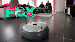 A robot vacuum cleaner cleans a tiled floor while people sit on couches in the background.