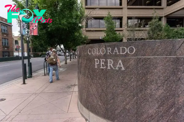 A person walking on a sidewalk past a large stone sign that reads "Colorado PERA," with buildings and trees in the background.