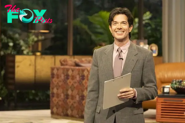 John Mulaney, holding a clipboard and wearing a suit, smiles on stage in Everybody’s in L.A.