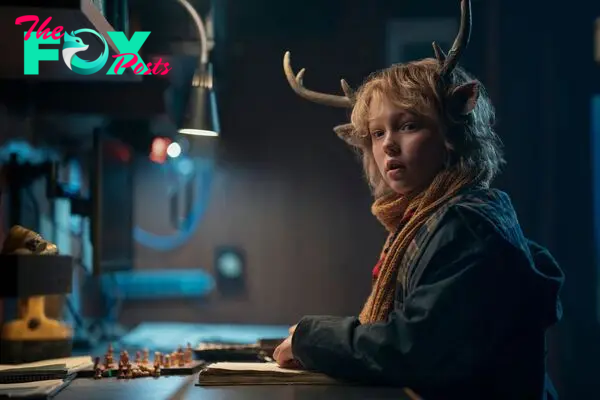 Christian Convery as Gus looks left at the camera while sitting at a desk in Sweet Tooth X3. Gus is a little boy who is dressed warmly, has a shaggy blonde mop, and the ears and antlers of a deer.