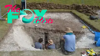 In this image we see a large square piece of grass that has been dug up to reveal a 10,000-year-old pit. Inside the pit are 2 archaeologists taking samples. There are 2 more archaeologists outside the pit, on opposite sides, taking measurements. Several tools are strewn about the site.
