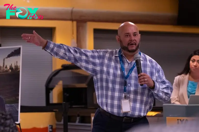 Joe Salazar, wearing a plaid shirt stands and speaks into a microphone, gesturing with one arm extended. A woman and a display board are visible in the background.