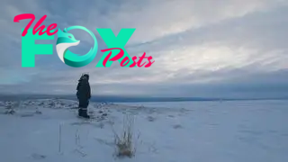 A man in winter clothing stands in an Arctic field with a few pieces of grass poking through the snow