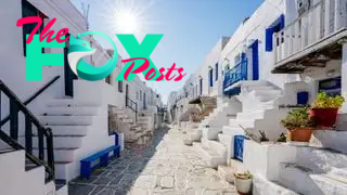 Image of a street in Greece lined with white square houses with blue accents. The sun is shining high in the sky.