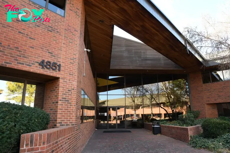 Brick building exterior with the number 4851 displayed. The entrance features large glass windows and an overhanging wooden structure. Bushes and a small brick wall surround the area near the doorway.