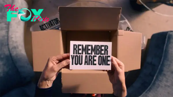 A person holds a card with the text &quot;REMEMBER YOU ARE ONE&quot; over an open cardboard box