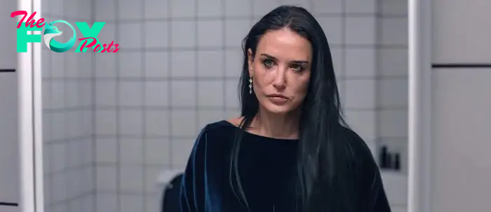 Demi Moore in a melancholy scene, looking directly at the camera, with long, straight hair and wearing a simple dark top. Background is a tiled wall