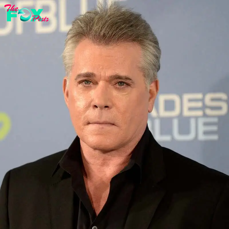 Ray Liotta at a "#ShadesOfBlue" event wearing a black suit jacket and shirt, posing in front of a backdrop with event branding and text