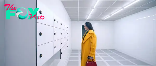 Woman in a stylish trench coat looks at minimalist wall storage units in a modern, brightly lit room. She holds a handbag. Names are unknown