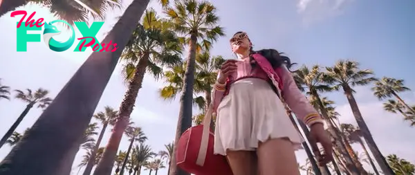 A person wearing sunglasses, a pink jacket, and a white skirt is walking among tall palm trees on a sunny day, holding a drink and bag. Names unknown