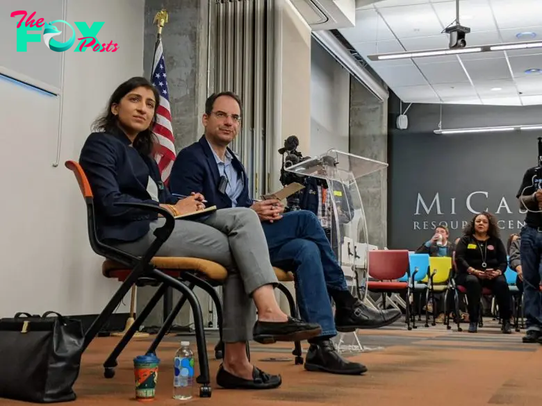 Lina Khan and Phil Weiser are seated on a stage in a conference room, discussing the Kroger-Albertsons merge as they face the audience. An American flag and various chairs are visible in the background, setting the scene for this significant industry milestone.