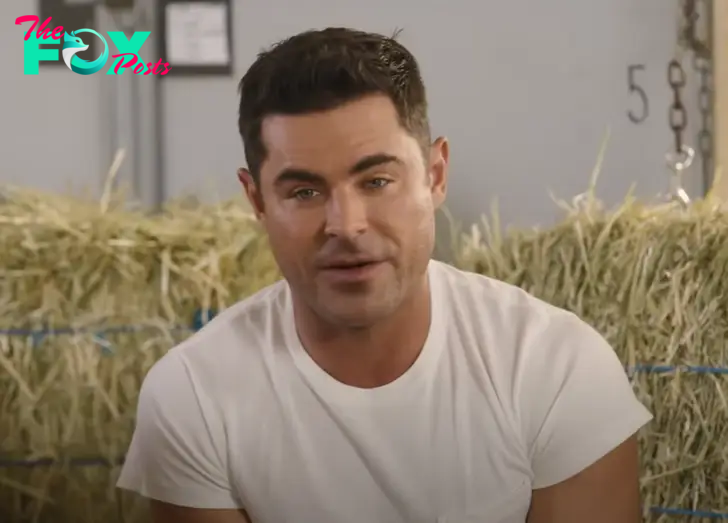 Close up of Zac Efron in a white shirt, stacks of hay in the background.