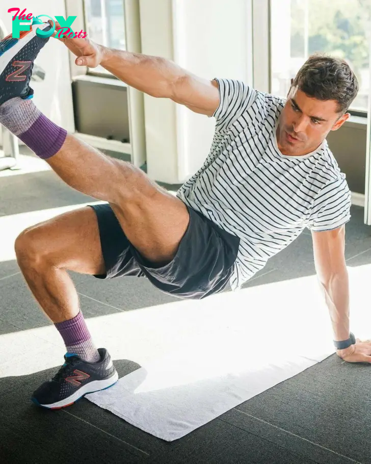 Zac Efron raising his feet and touching it with his hand, wearing white tshirt and black shorts.