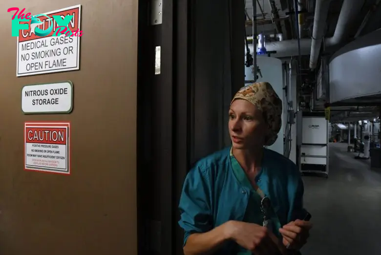 A person in scrubs and a cap stands beside a door with caution signs warning about medical gases, no smoking, and nitrous oxide storage in a dimly lit industrial hallway.