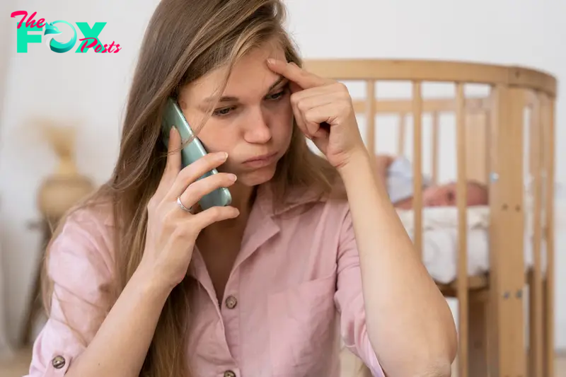 A frustrated woman talking on the phone with her baby lying in a cot behind her | Source: Freepik