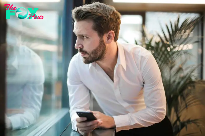 A man looking out the window, phone in hand | Source: Pexels