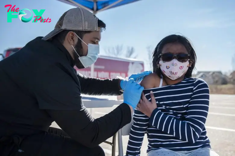 A healthcare worker wearing gloves and a mask administers a COVID vaccine to a child with glasses and a mask, outside in a tent-covered area. The child is in a striped shirt and is looking away.