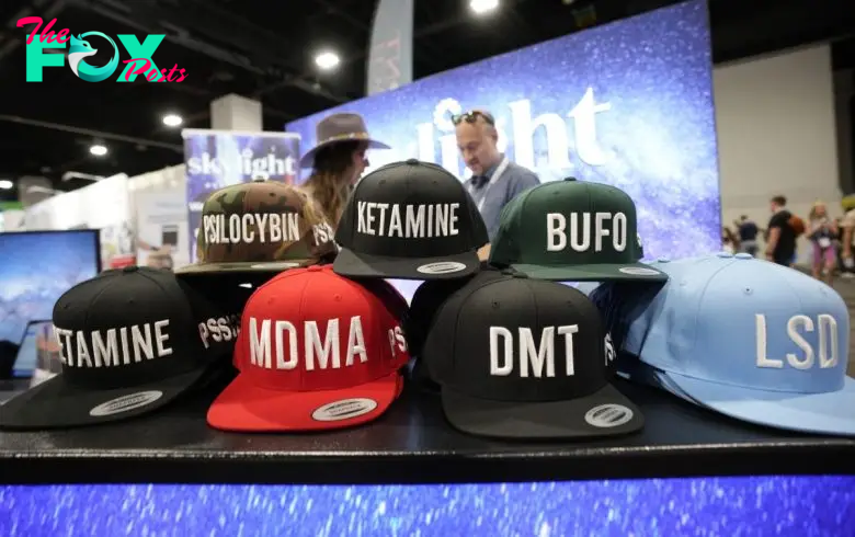 A display of hats with words like "PSILOCYBIN," "KETAMINE," and "LSD" at an exhibit booth.